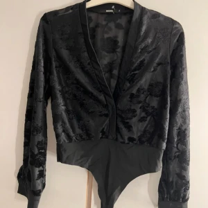 Svart blommig bodysuit  - Snygg svart bodysuit från BIK BOK med blommigt sammetmönster och djup v-ringning. Långärmad modell med transparent mesh och täckt nederdel. Perfekt för dig som vill ha en statement-top med coola detaljer och elegant vibe.