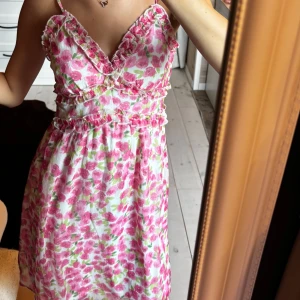 Blommig kortklänning med volang - Supersöt kortklänning från Vero Moda med rosa och grönt blommönster på vit botten. Klänningen har smala axelband, v-ringning och volangdetaljer längs byst och midja. Perfekt för sommarens alla tillfällen!