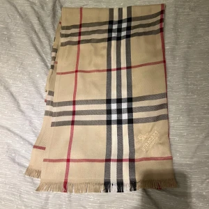 Beige rutig halsduk från Burberry - Klassisk beige halsduk från Burberry med ikoniskt rutigt mönster i svart, vitt och rött. Halsduken har fransar i kanterna och broderad Burberry London-logga. Perfekt accessoar för att lyfta din outfit.