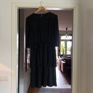 Svart prickig långklänning med knyt - Vintage svart medellång klänning med vita små prickar, trekvartsärmar med knyt vid ärmslut och markerad midja. Klänningen har ett luftigt fall och volang i kjolen. Strl: 44