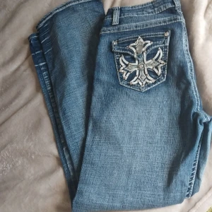 Blåa Ed Hardy jeans med detaljer - Snygga blå jeans från Ed Hardy med raka ben och coola broderade bakfickor med silvriga nitar och korsdetaljer. Jeansen har klassisk femficksdesign, normal passform och är tillverkade i mjukt bomullsjeansmaterial.