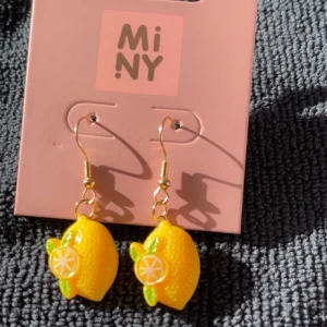 Citronörhängen från MiNY - Supergulliga örhängen från MiNY med hängande citroner i starkt gul färg och små gröna blad. Perfekt för dig som vill sticka ut med ett lekfullt och färgglatt smycke. Krokarna är i guldfärg och citronerna har en 3D-effekt.