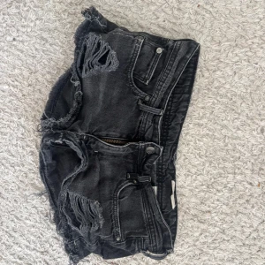 Svarta slitna jeansshorts - Riktigt snygga svarta jeansshorts med slitna detaljer och fransig kant. Klassisk femficksmodell med dragkedja och knapp framtill. Perfekta för en edgy streetstil och passar till allt.