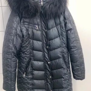 Svart dunjacka Rockandblue pälsdetalj - Snygg svart dunjacka från Rockandblue med stor fluffig päls runt kragen. Jackan har quiltad design, dragkedjor på fickor och bröst, samt tryckknappar framtill. Perfekt för kalla dagar och ger en cool vibe till vinterlooken.