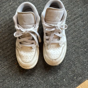 Vita Nike Jordan 1 sneakers - Snygga vita Nike Jordan 1 sneakers med klassisk siluett och perforerad tå. Skorna har snörning och rund tå, samt den ikoniska Jumpman-loggan på plösen. Tillverkade i skinn med vit sula och detaljer. Perfekta för dig som gillar streetstyle.