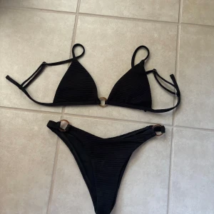 Svart ribbad bikini med ringdetaljer - Snygg svart bikini med ribbad struktur och trendiga ringdetaljer i brunt på både över- och underdel. Triangelformad topp med smala axelband och matchande trosa med högt skuren passform. Perfekt för stranden eller poolen i sommar!