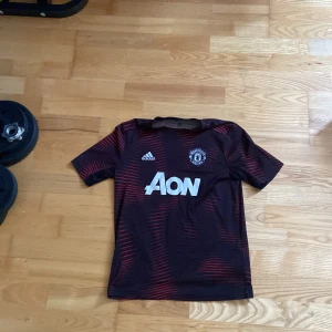 Fotbollströja united - Snygg Manchester United fotbollströja från Adidas med Aon-logga på bröstet. Tröjan är svart med röda ränder och har klubbmärket på vänster sida. Tillverkad i lätt och ventilerande material, perfekt för träning eller match.