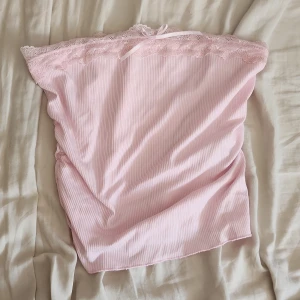 Ljusrosa ribbad tubtopp med spets - Supersöt ljusrosa tubtopp med ribbad struktur och spetskant upptill. Toppen har en liten rosett framtill och är axelbandslös, perfekt för en söt och feminin look. Mjuk och stretchig, passar perfekt till sommarens outfits.