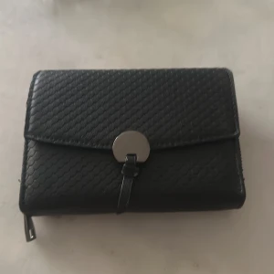 Svart plånbok med struktur och dragkedja - Snygg svart plånbok i skinnimitation med strukturerad yta och silverfärgat spänne framtill. Plånboken har flera kortfack, fönster för ID och ett myntfack med dragkedja. Perfekt storlek för att få plats med allt du behöver.