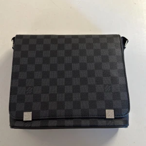 Louis Vuitton axelväska Damier Graphite - Snygg axelväska från Louis Vuitton i klassiska Damier Graphite-mönstret. Väskan är svart med rutigt mönster och har justerbar axelrem. Tillverkad i tåligt skinn och har silverfärgade metalldetaljer. Perfekt för dig som vill ha en stilren och lyxig väska.