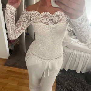 Vit offshoulder spets body - Superfin vit body i spets med offshoulder-modell och långa ärmar. Blusen har ett blommigt spetsmönster och sitter tight mot kroppen. Perfekt för dig som vill ha en feminin och elegant look.