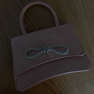 Rosa handväska med glittrig rosett - Söt liten rosa handväska i syntet med krokopräglad yta och ett glittrigt rosettdetalj framtill. Väskan har ett stadigt handtag och stilren form, perfekt för att lyfta din outfit med en touch av glamour.