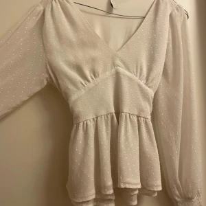 Vit blus från Amisu - Säljer en söt vit blus från Amisu med v-ringning och knytband i ryggen. Blusen har långa ärmar med elastiska muddar och ett diskret prickigt mönster. Perfekt för en stilren look.