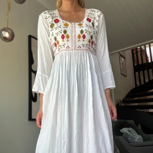 Vit långklänning med broderi - Vit tibetansk långklänning i bomull med färgglada broderade blommor i rött, gult och grönt över bysten. Funkar bra med neutrala underkläder/underkjol! Fler bilder finns och pris kan diskuteras.