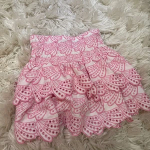 Rosa volangkjol från Kappahl - Supersöt kortkjol från Kappahl i vitt med rosa broderat mönster och flera volanger. Kjolen har bred resår i midjan för skön passform och ett lekfullt, somrigt uttryck. Perfekt för dig som gillar färg och detaljer! Helt ny me prislapp 
