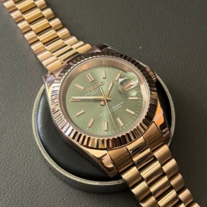 Seiko Datejust olive green presidential - Snygg Seiko Datejust med guldigt armband i metall och en stilren grön urtavla. Klockan har räfflad bezel, datumfönster med förstoringsglas och klassisk länkdesign. Perfekt accessoar för dig som gillar en lyxig och modern look.