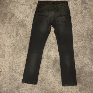 Svarta jeans från Nudie Jeans - Snygga mörk gråa jeans från Nudie Jeans med en klassisk femficksdesign med en diskret logga där bak. De är i väldigt bra skick då de bara har använts fyra gånger. Jeansen sitter slim och är perfekta för en stilren look. Priset är ej hugget i sten och går att diskutera.