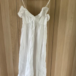 Vit långklänning med volang från H&M - Superfin vit långklänning från H&M med tunna axelband och volangdetaljer vid urringningen. Klänningen har en luftig passform och är tillverkad i bomull, perfekt för varma dagar. Enkel och stilren design som känns både romantisk och trendig.