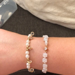 Två små söta armband i vit o guld - Säljer två söta armband, ett med vita pärlor, guldiga detaljer och små glittriga stenar, och ett med vita blommor, guldiga detaljer och en fyrkantig kristall. 