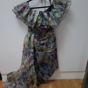 Blommig långklänning med volang - Superfin långklänning med blommigt mönster i lila, grönt, blått och gult. Klänningen har stora volanger upptill och en omlottliknande kjol. Tunt, flowy material som känns lyxigt och somrigt. Perfekt för dig som vill sticka ut med en romantisk look.