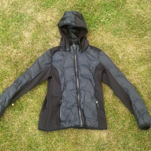 Svart pufferjacka från JH Collection - Svart pufferjacka från JH Collection med huva och dragkedja framtill. Jackan har quiltade partier och är fodrad med mjuk fleece på insidan. Perfekt för kyliga dagar och har en sportig look med snygga detaljer och fickor med dragkedja.