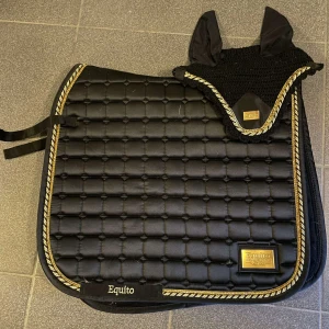 Svart Equito huva med guld-detaljer och ett matchande shcabrak  - Snygg svart huva från Equito med broderade öron där det står 'enjoy the ride' och 'sit back' i guld. Huva i virkad stil med glittrig guld- och svart kant. Perfekt för ridning och matchar schabrak i samma stil.