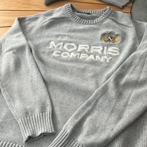 Ljusgrå stickad tröja Morris Company - Snygg ljusgrå stickad tröja från Morris Company med broderad logga och emblem på bröstet. Tröjan har rund halsringning, ribbade muddar och är långärmad. Perfekt för dig som gillar stilren och klassisk look.