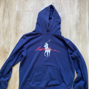 Marinblå hoodie från Polo Ralph Lauren - Snygg marinblå hoodie från Polo Ralph Lauren med stor vit polospelare och röd signatur på bröstet. Klassisk känguruficka framtill, huva med snörning och mjukt bomullsmaterial. Ingen defekter alls! Skriv vid fler funderingar 🙌