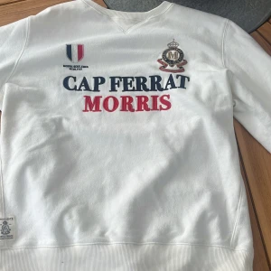 Vit sweatshirt från Morris, Cap Ferrat - Vit sweatshirt från Morris med tryck och broderade detaljer på bröstet och ärmen. Klassisk rund halsringning, ribbade muddar och normal passform. Snyggt tryck i marinblått och rött framtill samt siffra på ärmen. Perfekt för en clean och sportig look.