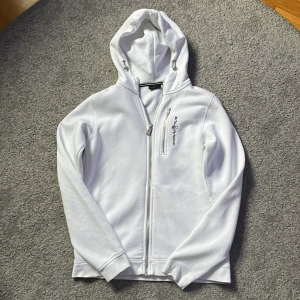 Vit Sail Racing hoodiejacka 176 - Vit hoodiejacka från Sail Racing med dragkedja framtill och på bröstfickan. Jackan har broderad logga, huva med snörning och är tillverkad i mjukt material med fleecefoder på insidan. Bra skick, använd runt 4-5 gånger.