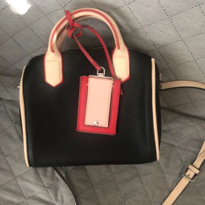 Svart handväska från Parfois - Stilren svart handväska från Parfois med kontrasterande beige handtag och detaljer samt en cool tag i rött och ljusrosa. Väskan har både handtag och axelrem, dragkedja upptill och flera fack inuti. Perfekt storlek för det viktigaste och snygg att bära till alla outfits.