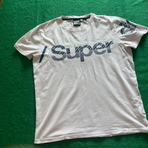 Rosa Superdry t-shirt med blått tryck - Rosa t-shirt från Superdry med stort blått 'Super' tryck och japanska tecken på bröstet. Klassisk passform med rund hals och korta ärmar. Snygg detalj med blått tryck även på ena ärmen. Perfekt för en avslappnad streetstyle.