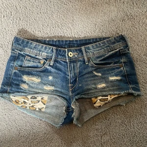 Blå jeansshorts med leoparddetaljer - Snygga blå jeansshorts från Denim med slitna detaljer och råa kanter. Insidan av shortsen har coola leopardmönstrade lappar som syns genom de trasiga partierna framtill. Klassisk femficksmodell och låg midja. Midjemått: 37cm🤗