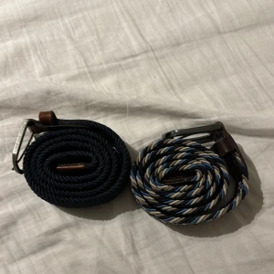 Två flätade bälten med metallspänne - Säljer två snygga flätade bälten med metallspänne. Det ena är enfärgat mörkblått och det andra har ett randigt mönster i blått, beige och vitt. Båda bältena har bruna läderdetaljer vid spännet. Perfekta för att ge din outfit en stilren touch! För både kan du köpa för 200