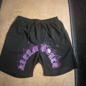 Svarta shorts med lila y2k tryck - Svarta shorts med elastisk midja och snörning. De har ett coolt lila tryck med texten 'Dream State' i eldliknande stil. Perfekta för en avslappnad stil.