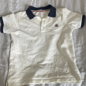 Vit pikétröja med mörkblå detaljer - Säljer en stilren vit pikétröja från Moncler med mörkblå krage och ärmslut. Tröjan har en broderad logga på bröstet och knappar framtill. Perfekt för en avslappnad men snygg look.