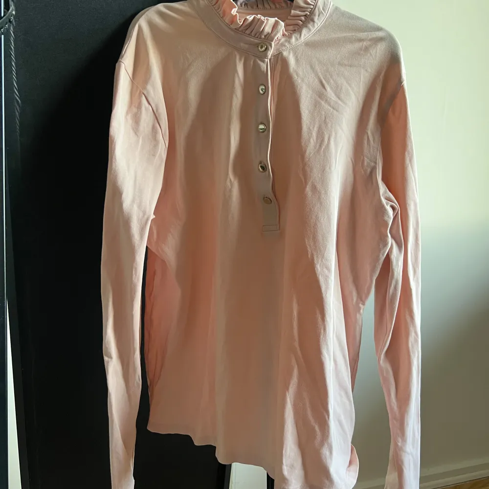 Säljer en ljusrosa långärmad topp från Holly & Whyte by Lindex. Toppen har en volangdetalj vid halsen och knappar framtill. Perfekt för en stilren look.. Yläosat.