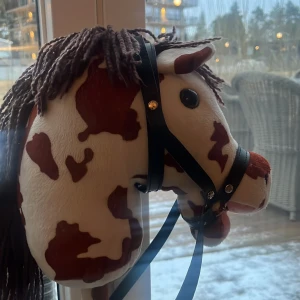 Käpphäst med brunt och vitt mönster - Charmig käpphäst med ett brunt och vitt fläckigt mönster. Hästen har en man av brunt garn och en svart träns. Perfekt för lek och äventyr! 