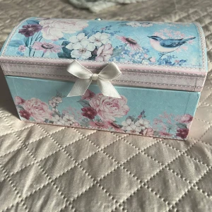 Blommigt smyckeskrin med fågelmotiv - Charmigt smyckeskrin med blommigt mönster och fågelmotiv. Skrinet har en ljusblå bas med rosa och vita blommor samt en söt rosett på framsidan. Perfekt för att förvara dina smycken och små skatter.