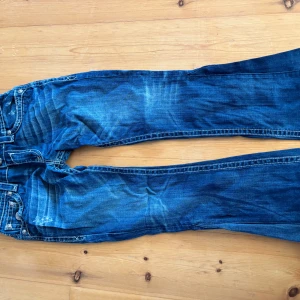 Blå jeans från Rock Revival - Säljer ett par snygga blå jeans från Rock Revival med coola slitningar och broderade detaljer på bakfickorna. Jeansen har en klassisk femficksdesign och en bekväm passform.