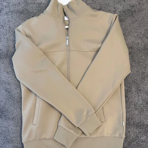 Beige tröja från Les Deux - Snygg beige jacka från Les Deux med dragkedja framtill och broderad logga på bröstet. Köpte denna för 1199kr och har endast använt en gång, men rensar garderob på gammla kläder så inget fel med den