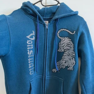 Rhinestone hoodie i färgen blå - Snygg blå hoodie från Vansydical med en cool tigerdesign i strass på framsidan. Tröjan har dragkedja och justerbara snören i huvan. Perfekt för en avslappnad stil.