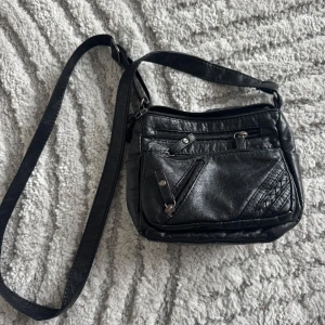 Svart axelväska i skinn - Snygg svart axelväska i skinn med flera dragkedjefickor för praktisk förvaring. Väskan har en justerbar axelrem och stilrena detaljer som ger en modern look. Perfekt för att bära dina nödvändigheter med stil.Den här väskan kommer även från 2000-talet.