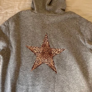 Grå hoodie med leopardstjärna - Säljer en grå hoodie med en stor leopardmönstrad stjärna på ryggen. Tröjan har en klassisk känguruficka och ribbade muddar vid ärmslut och nederkant. Perfekt för en avslappnad stil!