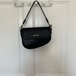 Svart axelväska från Valentino - Snygg svart axelväska från Valentino i skinn med guldfärgade detaljer. Väskan har en stilren design med justerbar axelrem och logotyp framtill. Perfekt för att ge en touch av lyx till din outfit.
