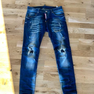 Blå jeans med slitningar från Dsquared - Snygga blå jeans från Dsquared med coola slitningar och färgstänk. De har en klassisk femficksdesign och en knappgylf. Skriv ifall du har har minsta fundering!