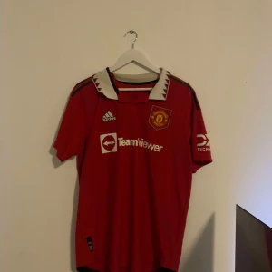 Röd fotbollströja från Adidas authentic - Säljer en röd fotbollströja från Adidas med Manchester Uniteds emblem på bröstet. Tröjan har korta ärmar och en vit krage med svarta detaljer. På baksidan finns namnet 'R. Varane' och nummer 19 tryckt i vitt. Storlek L