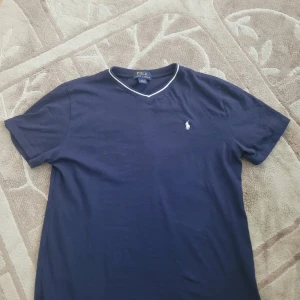 Mörkblå t-shirt från Ralph Lauren - Säljer en snygg mörkblå t-shirt från Ralph Lauren med v-ringning och en liten broderad logga på bröstet. T-shirten är tillverkad i 100% bomull och har korta ärmar. Använd bara ett par gånger. Jag kan frakta inom 24 timmar. Fråga gärna frågor.