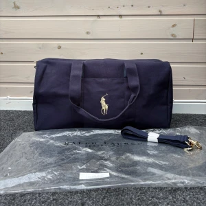 Mörkblå dufflebag/väska från Ralph Lauren - Snygg mörkblå dufflebag från Ralph Lauren med ett klassiskt broderat logotypmotiv. Väskan har en praktisk dragkedja i guld och en avtagbar axelrem. Perfekt för resor eller gymmet. Denna väska är i samband med Ralph lauren parfym då man fick med denna väska när man köpte en parfym, går därför inte få tag på, priset är ej hugget i sten så kom med förslag!, 