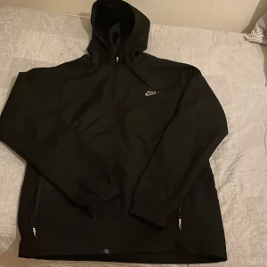 Svart vindjacka från Nike - Säljer en stilren svart vindjacka från Nike med dragkedja och huva. Jackan har långa ärmar och två praktiska fickor med dragkedja framtill. Perfekt för blåsiga dagar. Pris kan diskuteras!!